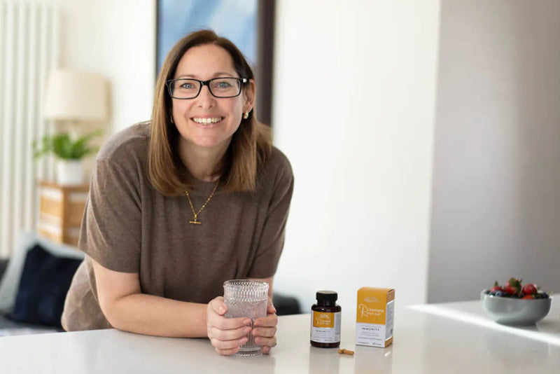 Woman standing at a table with a glass of water, supplement bottle and box on a white surface.