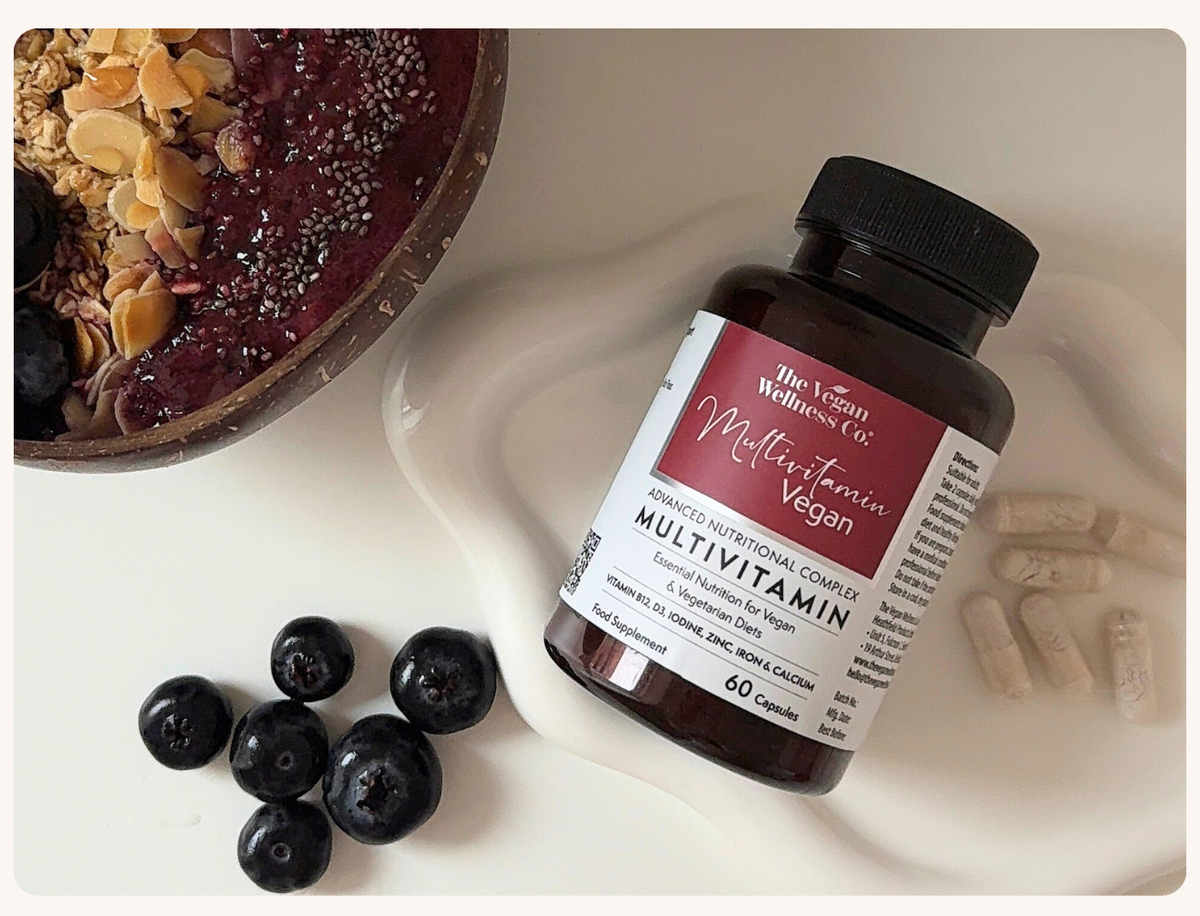 A bottle of Wellness Co. Multivitamin Vegan capsules arranged on a white surface with blueberries, granola, and a smoothie bowl in the background.